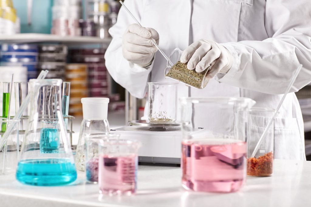 Equipped doctor making dermatology products in laboratory. Researcher wearing white gown and gloves putting samples of herbs in jar on scales while working on new formula of herbal organic goods